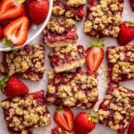 Homemade strawberry oatmeal bars with a buttery oat crumble topping arranged on a table with fresh sliced strawberries.