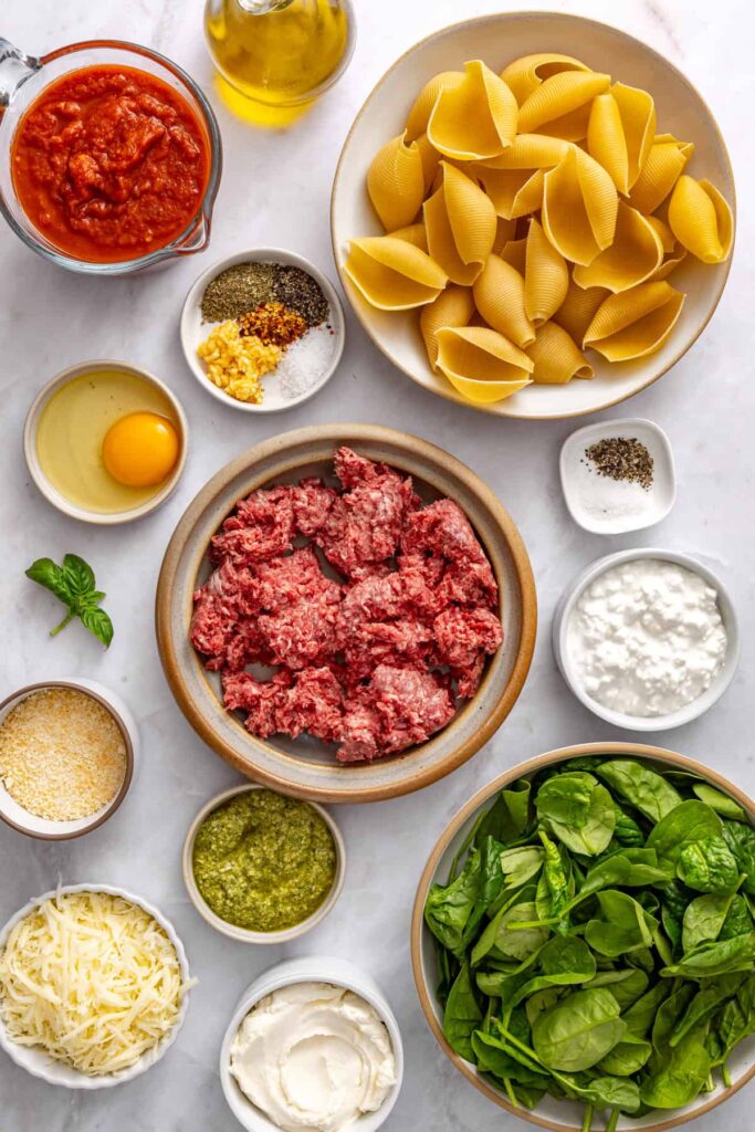 Overhead shot of ingredients for stuffed shells including pasta shells, ground beef, spinach, ricotta, pesto, and marinara sauce