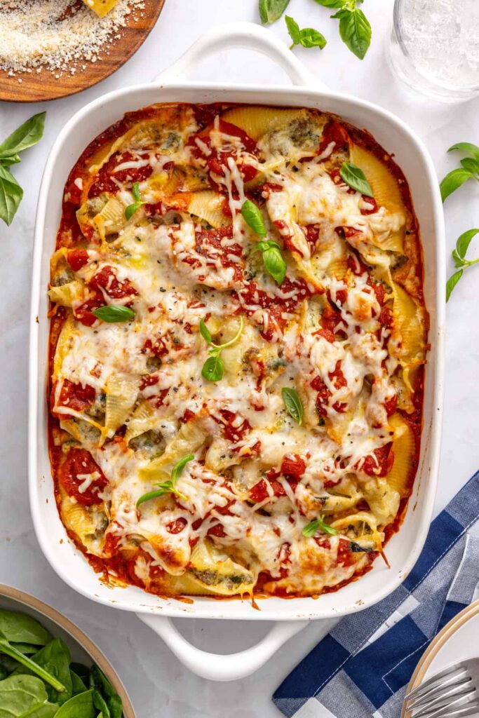 Overhead shot of baked stuffed shells with pesto beef, spinach, and whipped ricotta in a baking dish garnished with fresh basil and olive oil on a styled table
