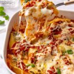 Close-up side angle of baked pesto beef and spinach stuffed shells in a baking dish being lifted with a serving spoon, showing melted cheese pull and creamy ricotta filling with a styled table in the background