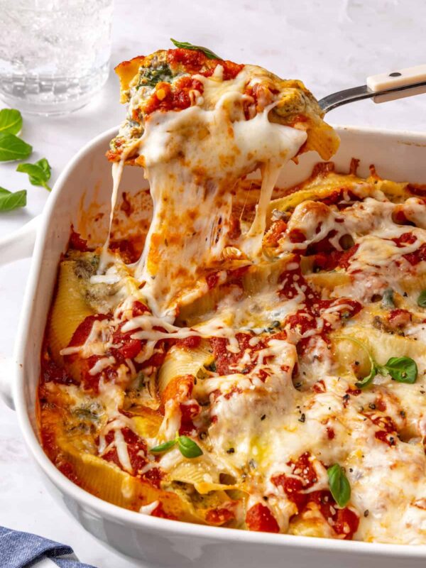 Close-up side angle of baked pesto beef and spinach stuffed shells in a baking dish being lifted with a serving spoon, showing melted cheese pull and creamy ricotta filling with a styled table in the background