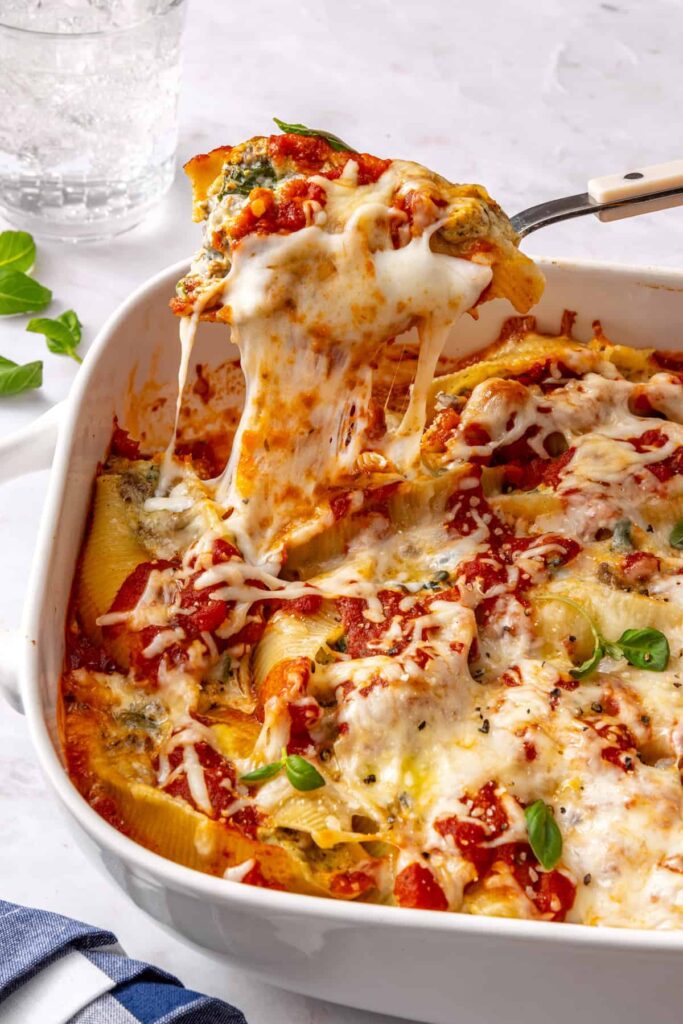 Close-up side angle of baked pesto beef and spinach stuffed shells in a baking dish being lifted with a serving spoon, showing melted cheese pull and creamy ricotta filling with a styled table in the background