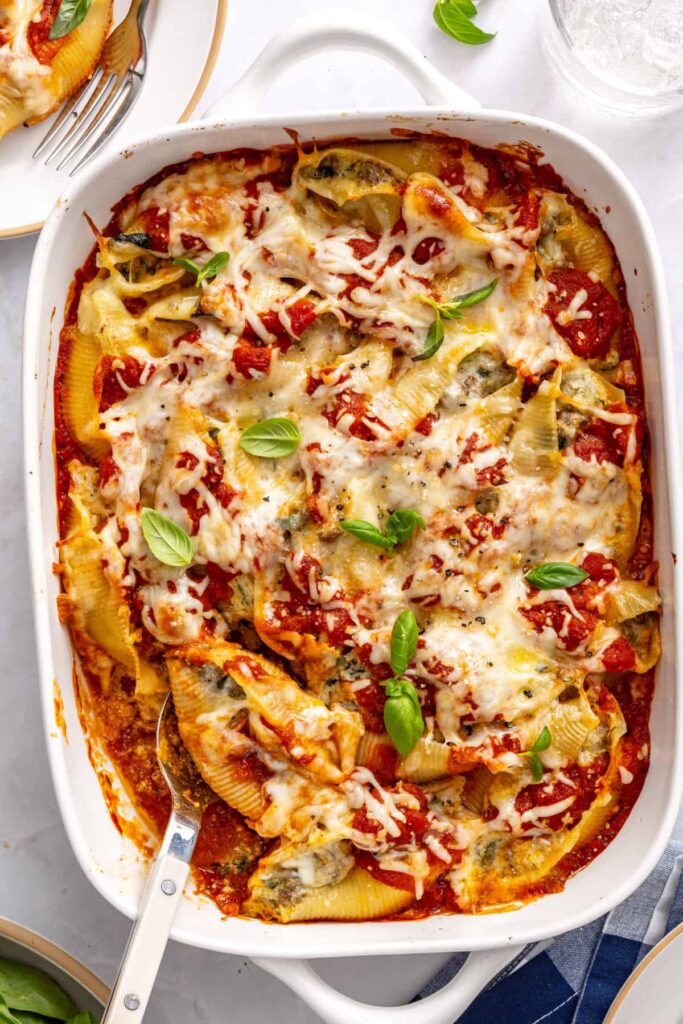 Overhead wide shot of baked stuffed shells in baking dish with serving spoon, plates, and drinks on styled table