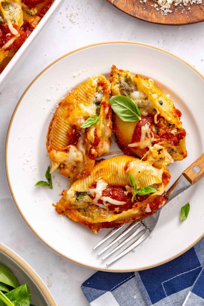 Serving of baked stuffed shells on plate with marinara and cheese, styled with tableware and checkered napkin
