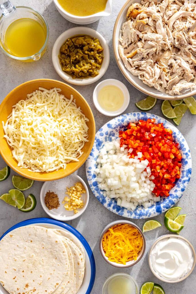 Ingredients for creamy green chicken enchiladas with salsa verde including shredded chicken, tortillas, cheeses, green enchilada sauce, vegetables, and lime
