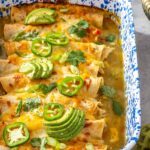 Creamy green chicken enchiladas with salsa verde baked in a speckled casserole dish and topped with avocado, jalapeños, and fresh cilantro
