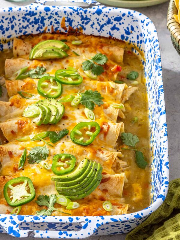 Creamy green chicken enchiladas with salsa verde baked in a speckled casserole dish and topped with avocado, jalapeños, and fresh cilantro