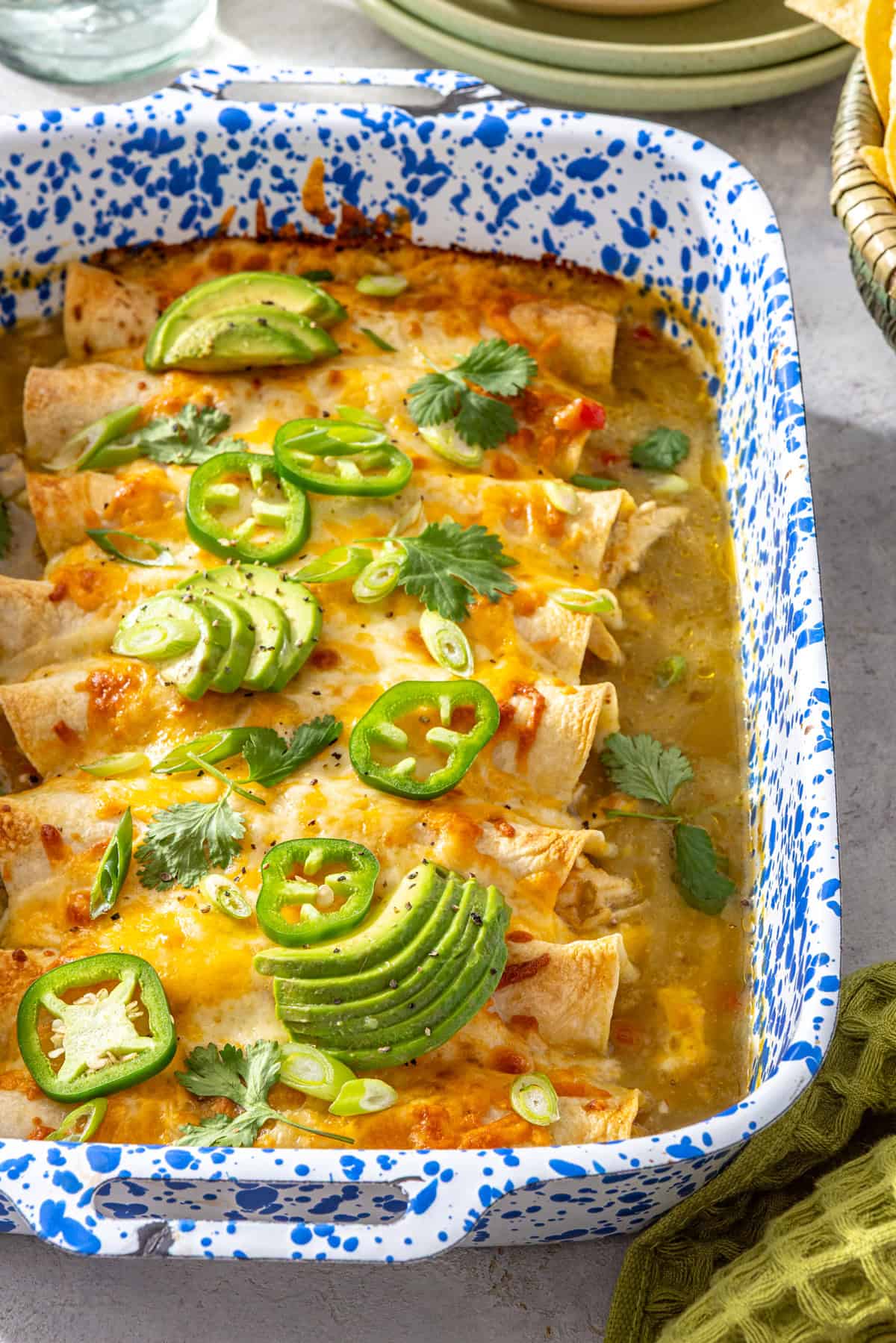 Creamy green chicken enchiladas with salsa verde baked in a speckled casserole dish and topped with avocado, jalapeños, and fresh cilantro