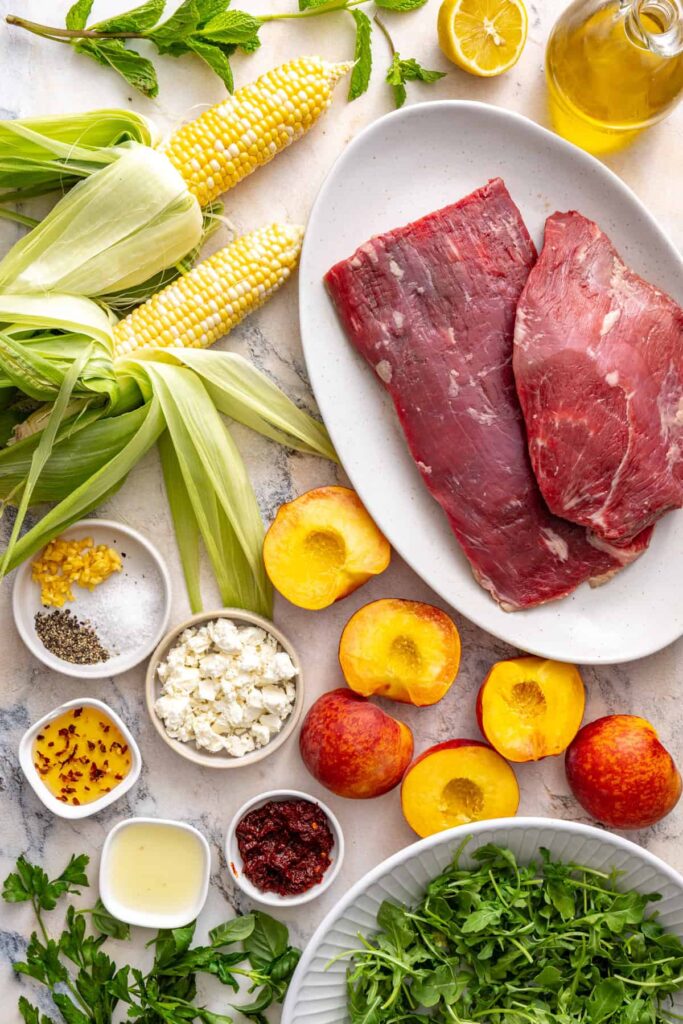 Ingredients for harissa steak salad including flank steak, peaches, corn, arugula, herbs, feta, and marinade ingredients
