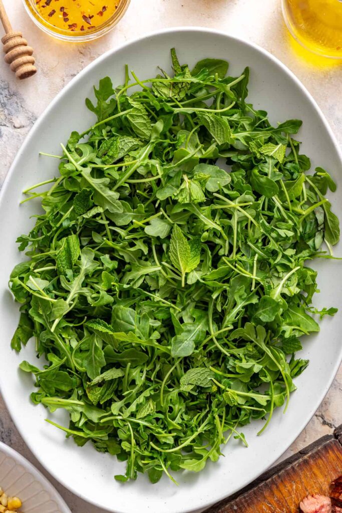 Fresh arugula in a salad bowl, ready to be topped with grilled steak, peaches, corn, and feta