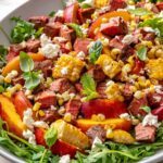 Harissa steak salad with charred peaches, grilled corn, arugula, feta, and fresh herbs served in a bowl