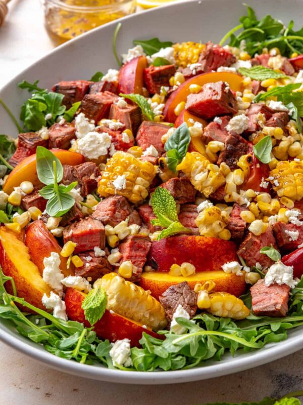 Harissa steak salad with charred peaches, grilled corn, arugula, feta, and fresh herbs served in a bowl