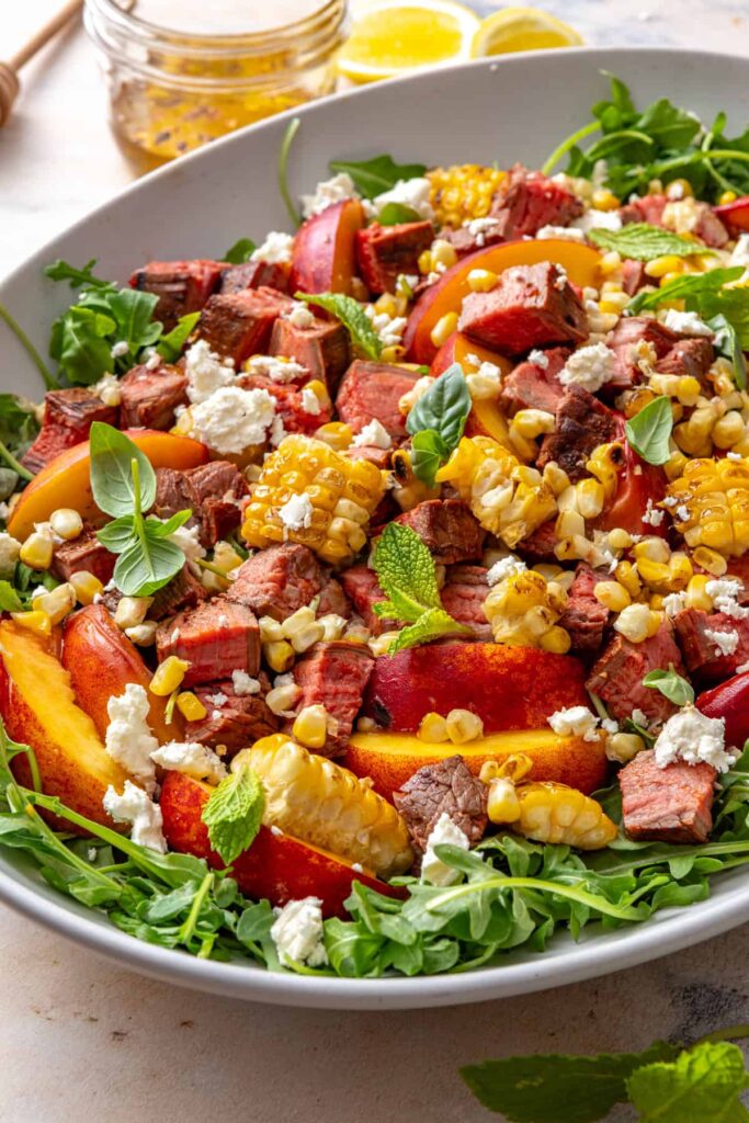 Harissa steak salad with charred peaches, grilled corn, arugula, feta, and fresh herbs served in a bowl