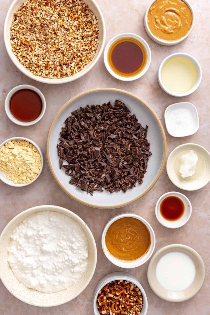 Overhead ingredients for frozen peanut butter pretzel bars including cottage cheese, peanut butter, gluten-free pretzels, dark chocolate, protein powder, maple syrup, and coconut oil.