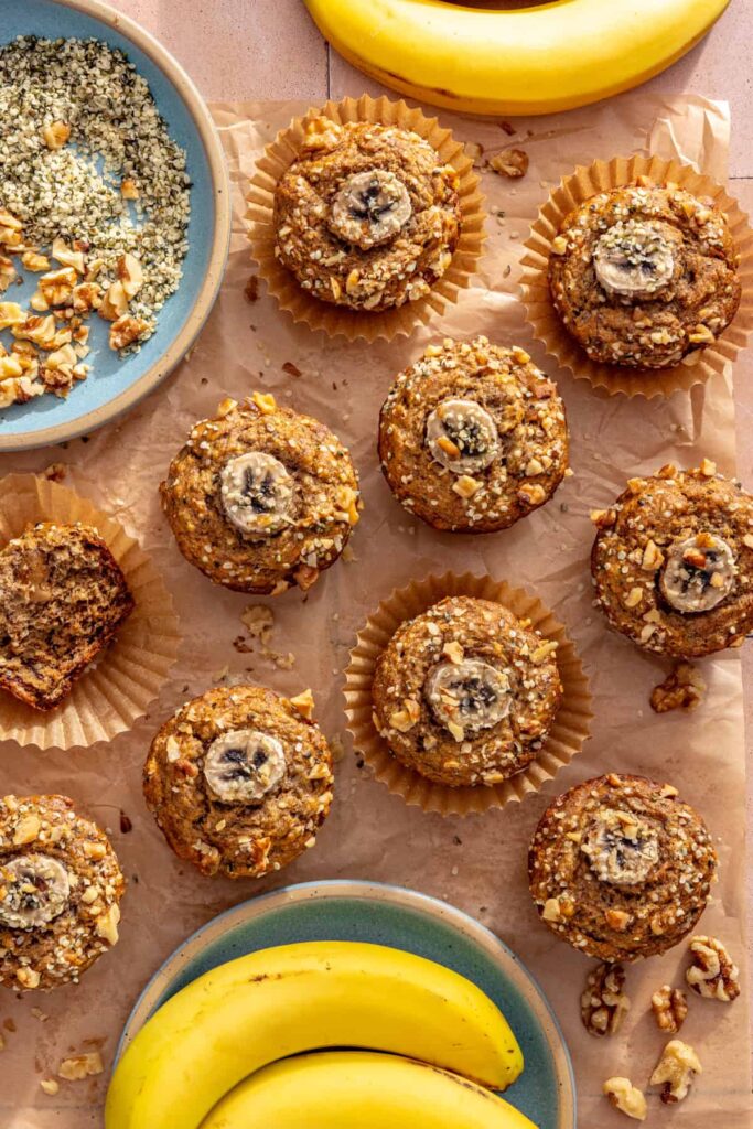 Toddler banana muffins wrapped individually on a table with fresh bananas, hemp seeds, and chopped walnuts