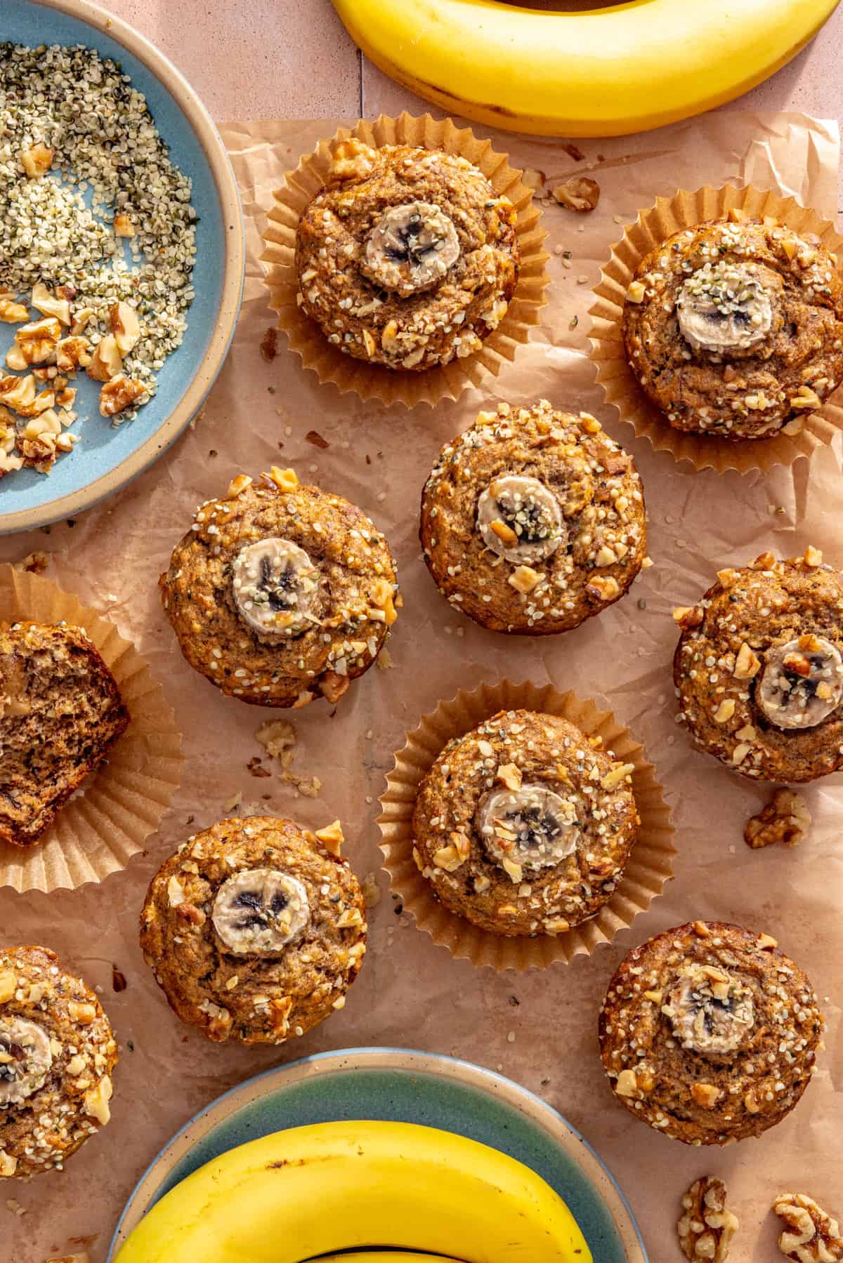 Toddler banana muffins wrapped individually on a styled table with fresh bananas, hemp seeds, and chopped walnuts