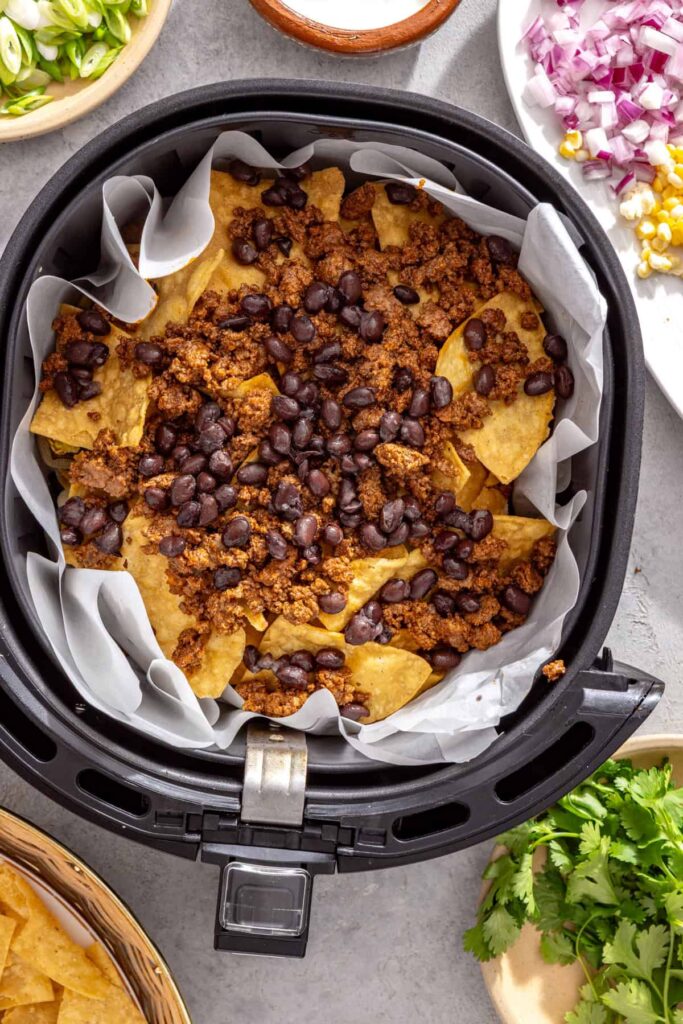 Air fryer nachos being layered with beef, beans, cheese, and colorful vegetables