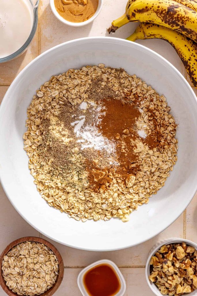 Rolled oats, cinnamon, nutmeg, flaxseed, hemp seeds, baking powder, and protein powder mixed in a bowl