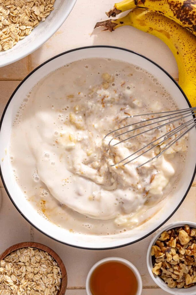 Whisking mashed bananas with almond milk, egg whites, maple syrup, vanilla, nut butter, and Greek yogurt