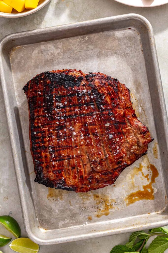 Grilled flank steak resting on a parchment paper-lined baking tray before slicing