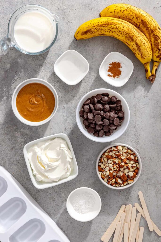 Ingredients for healthy homemade ice cream bars including bananas, Greek yogurt, peanut butter, milk, cinnamon, chocolate chips, coconut oil, chopped nuts, and flaky sea salt