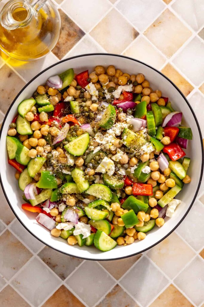 Chopped vegetables, chickpeas, capers, and feta added to a mixing bowl for chickpea salad