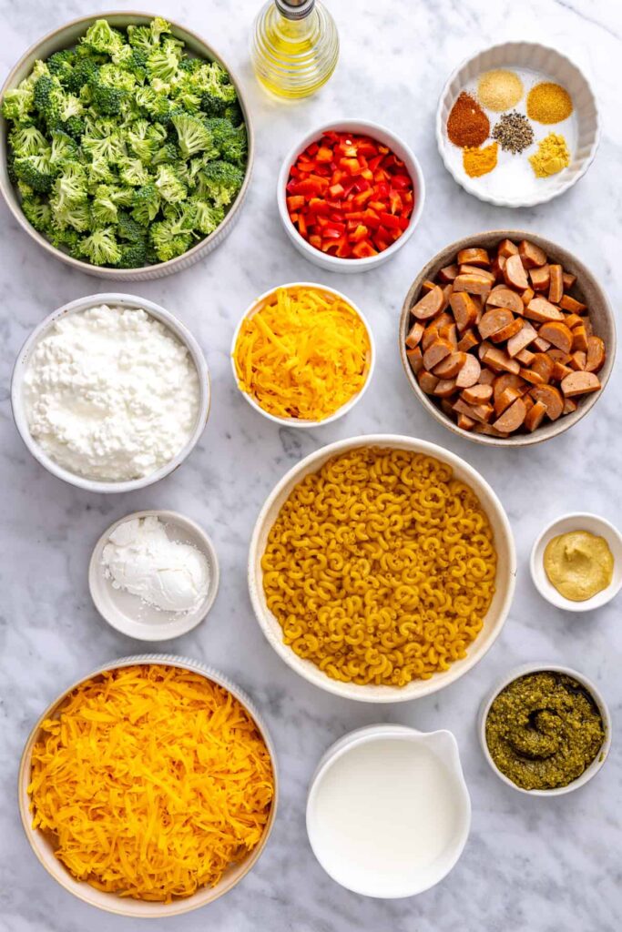 Ingredients for gluten-free broccoli cheddar pasta including cottage cheese, cheddar, broccoli, pasta, chicken sausage, red bell pepper and pesto