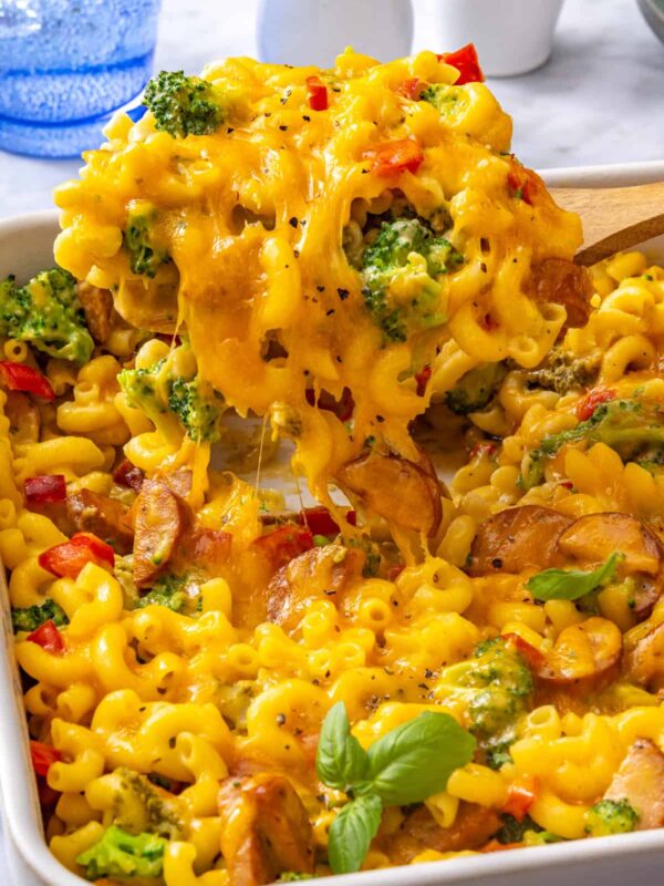 Close-up of creamy broccoli cheddar pasta with a spoon lifting cheesy pasta from the baking dish, showing melted cheddar, broccoli and chicken sausage