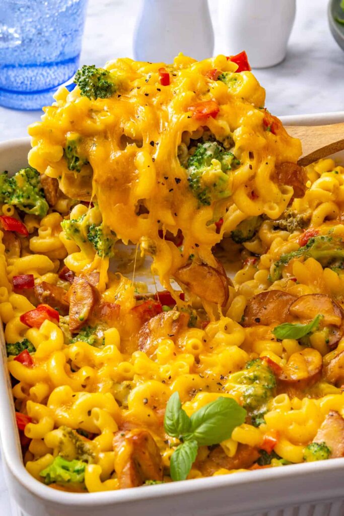 Close-up of creamy broccoli cheddar pasta with a spoon lifting cheesy pasta from the baking dish, showing melted cheddar, broccoli and chicken sausage