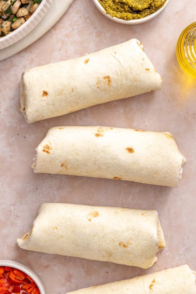 Three tightly rolled chicken pesto breakfast burritos ready to crisp in a skillet