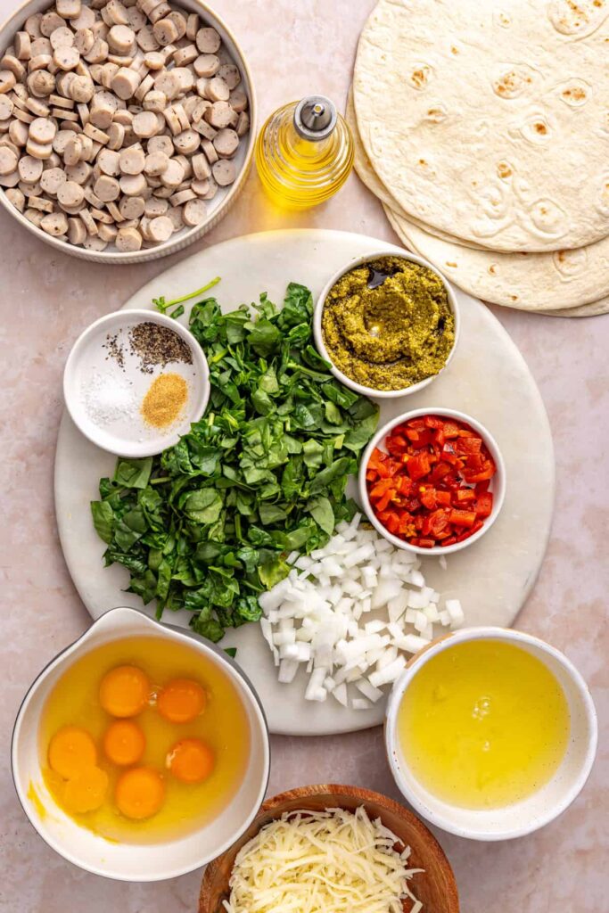 Ingredients for chicken pesto breakfast burritos, including chicken sausage, eggs, spinach, roasted red peppers, mozzarella, pesto and tortillas