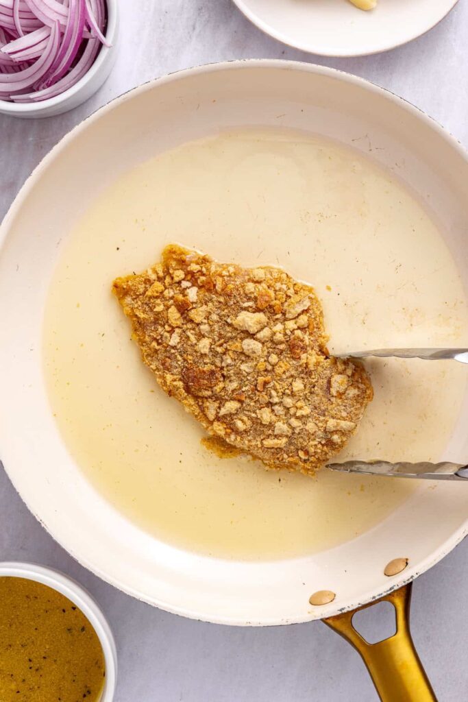 Chicken cutlet being flipped in a skillet to brown the second side during pan-frying.