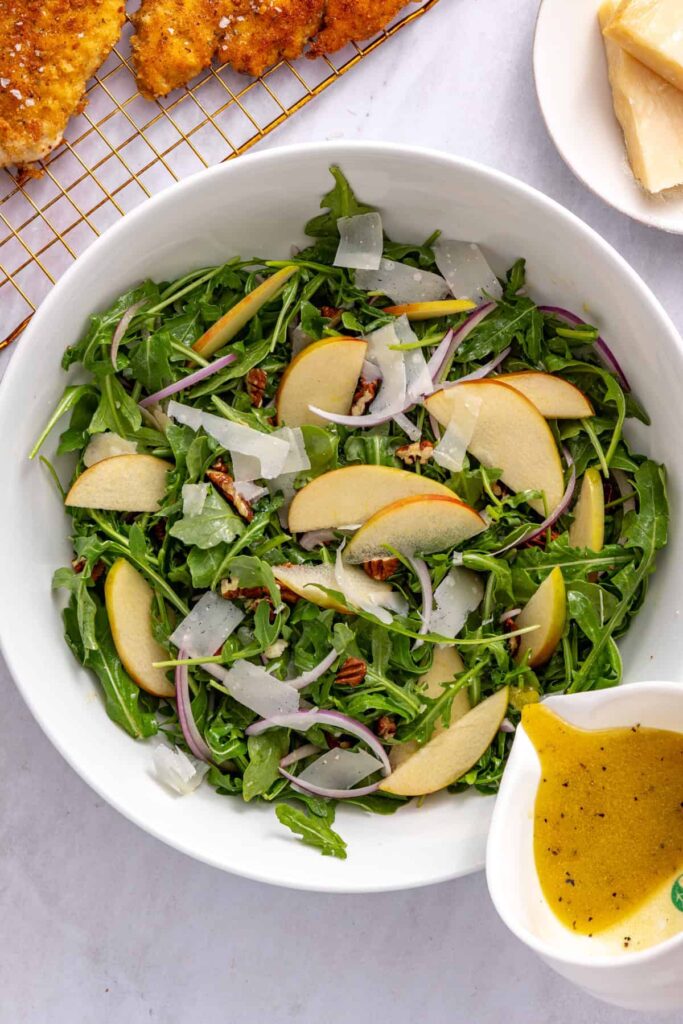 Maple Dijon vinaigrette being poured over fresh apple arugula salad with toasted pecans and sliced apple.
