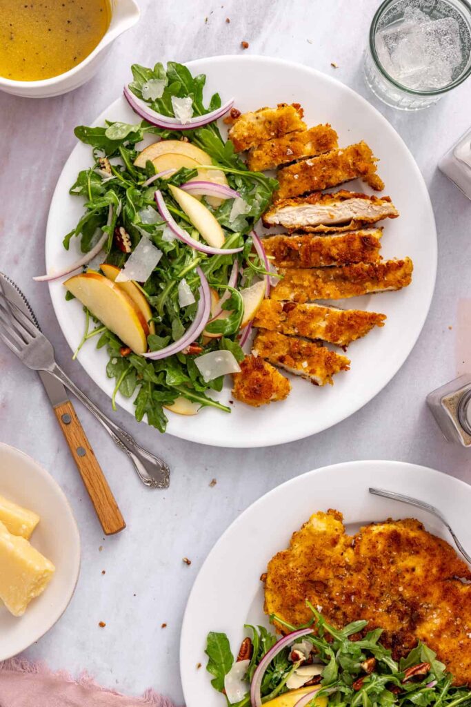 Overhead view of sliced golden chicken cutlets and fresh apple arugula salad with pecans on white plates at a styled table.