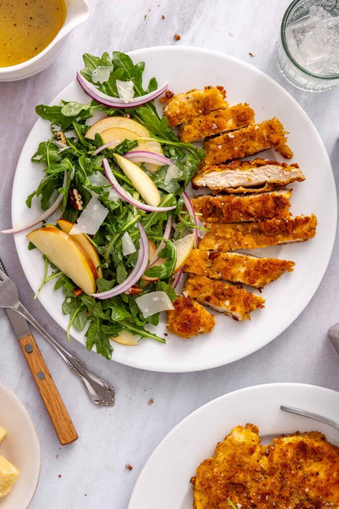 Crispy chicken cutlets with apple arugula salad with forks, knives, and water glasses.