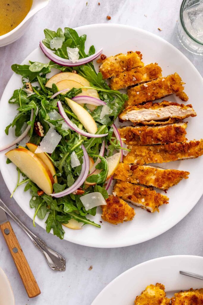 Close-up overhead view of crispy chicken cutlets served with apple arugula salad, shaved Parmesan, and toasted pecans.