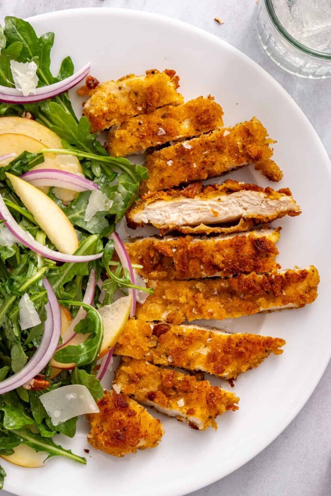 Close-up of sliced crispy chicken cutlets with a golden sourdough breadcrumb coating, served alongside apple arugula salad. Copy