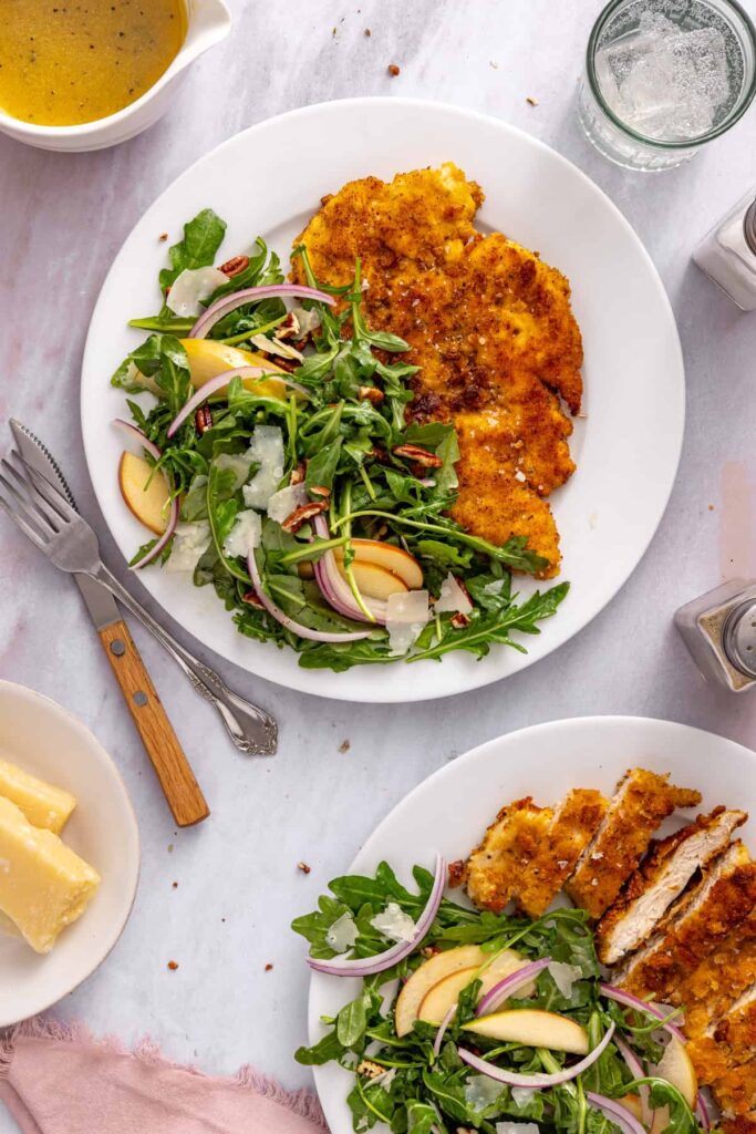 Crispy pan-fried chicken cutlets with a fresh Honeycrisp apple and arugula salad on a white plate, photographed from above.