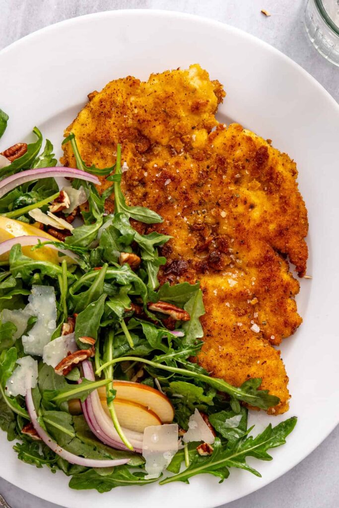 Golden pan-fried chicken cutlets sliced into strips on a white plate with fresh apple arugula salad on the side.