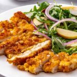 Close-up of crispy chicken cutlets with apple arugula salad and maple Dijon vinaigrette on a white plate.
