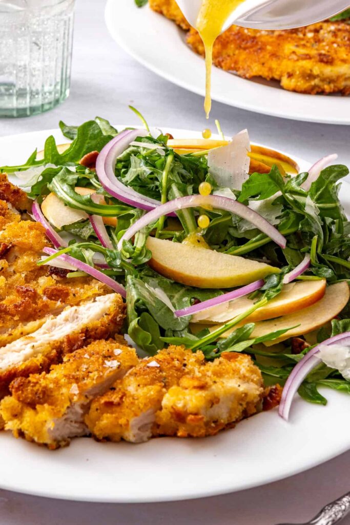 Maple Dijon vinaigrette drizzled over apple arugula salad served beside golden crispy chicken cutlets on a white plate.