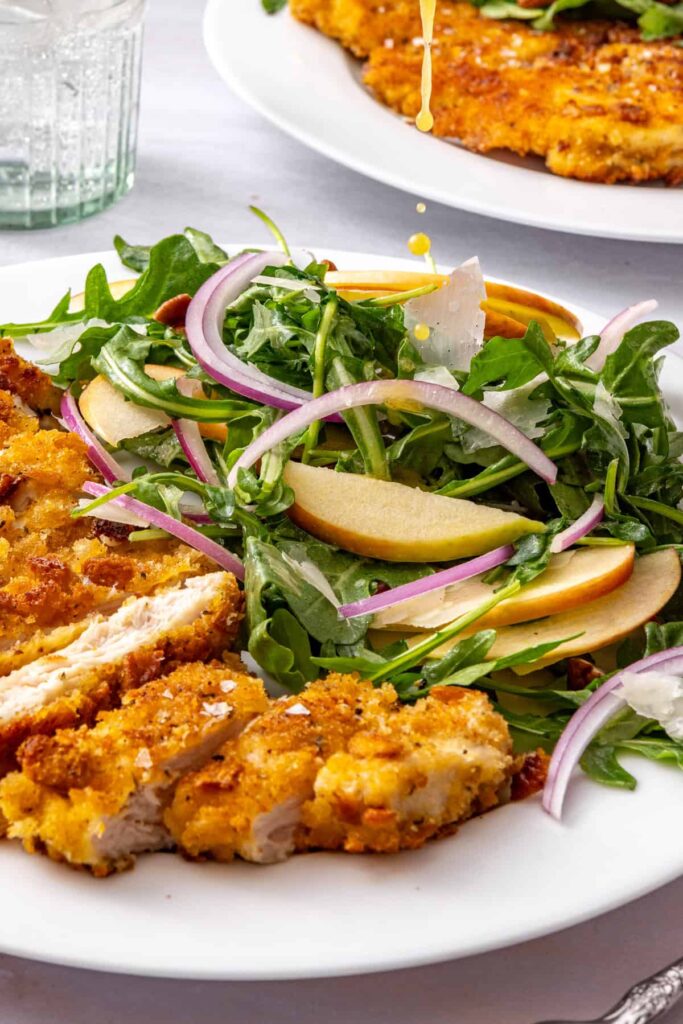 Close-up side view of golden maple Dijon dressing dripping onto fresh arugula and sliced apple beside crispy breaded chicken cutlets.