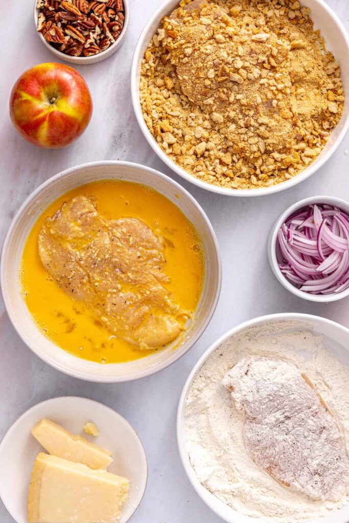 Three shallow bowls of flour, egg wash, and seasoned sourdough breadcrumbs prepared for breading chicken cutlets.