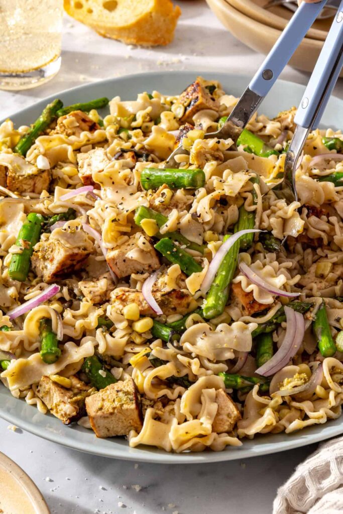 Serving spoons lifting chicken pesto pasta salad from a serving bowl