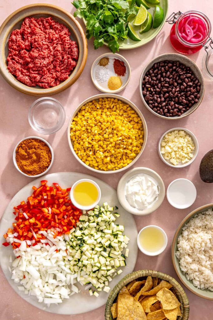 Ingredients for street corn taco skillet including ground beef, black beans, fire-roasted corn, zucchini, bell pepper, onion, Greek yogurt, cotija cheese, and lime