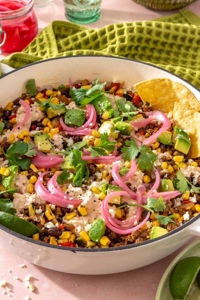 Side angle of finished street corn taco skillet garnished with pickled red onions, cotija, cilantro, lime, and tortilla chips