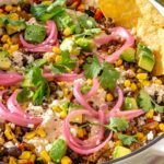 Close-up side angle of street corn taco skillet with tortilla chips dipping into the skillet, topped with pickled red onions, cotija, cilantro, and lime