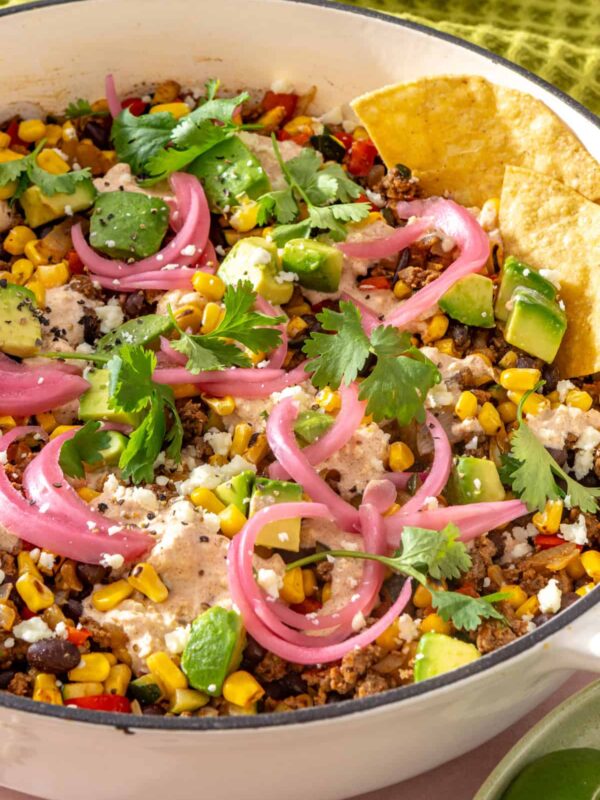Close-up side angle of street corn taco skillet with tortilla chips dipping into the skillet, topped with pickled red onions, cotija, cilantro, and lime