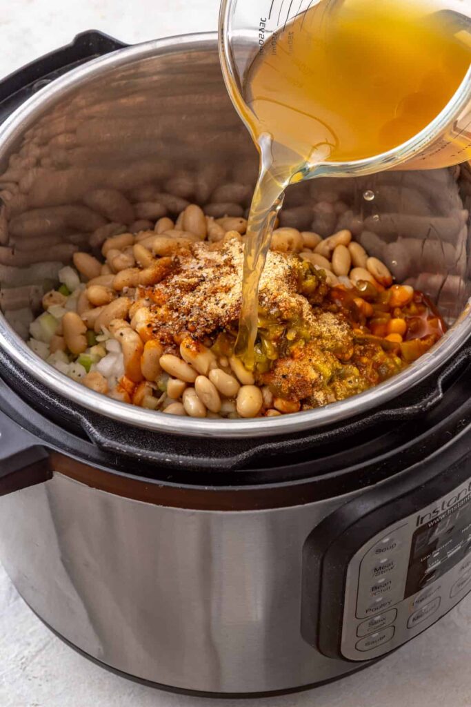 bone broth being poured into an Instant Pot on top of chicken thighs, cannellini beans and vegetables for a chicken chili recipe