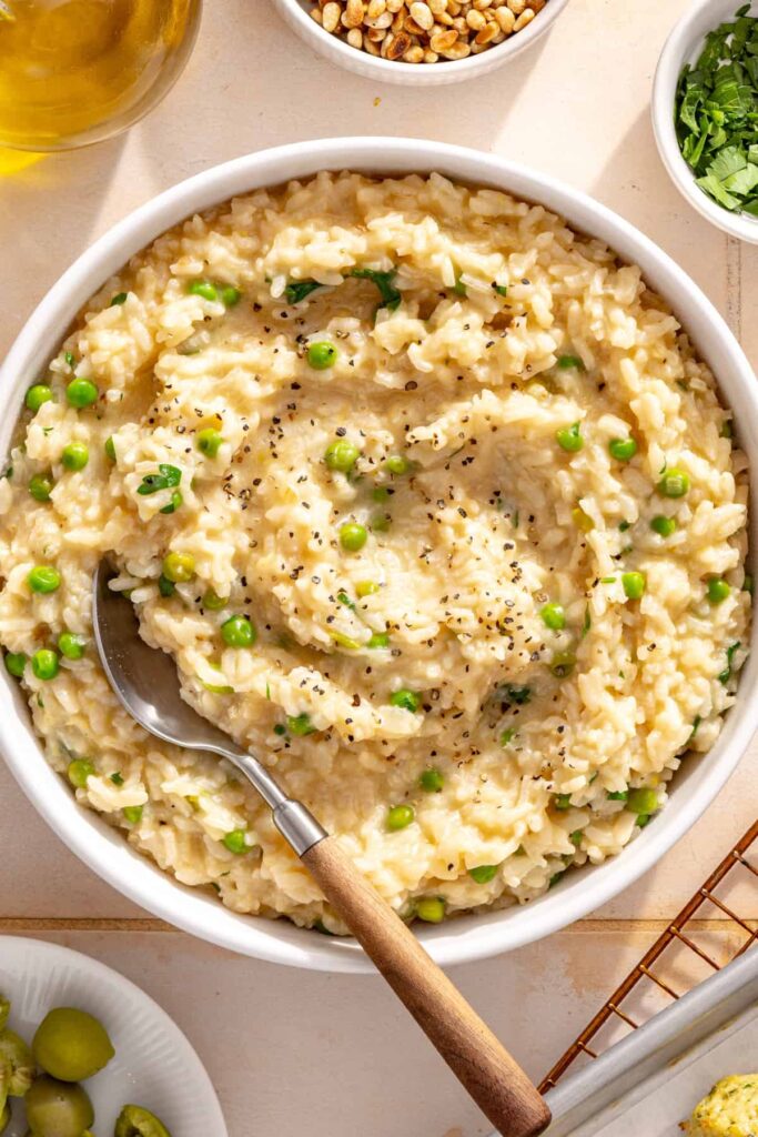 Bowl of creamy parmesan rice garnished with toasted pine nuts, green olives and fresh herbs